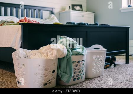 Three white plastic laundry baskets lined up on a beige carpet floor near a bed with a white and cream colored quilt Stock Photo