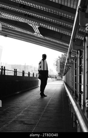Black & white image of silhouette of man standing in urban passageway with low roof, leading lines & vanishing point against bright sky. Stock Photo
