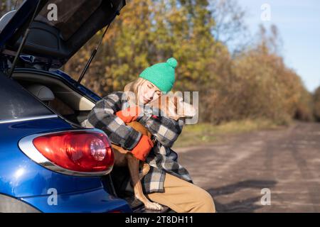 Middle aged woman in road trip with dog hugging pet sitting in car trunk during break on nature.  Stock Photo