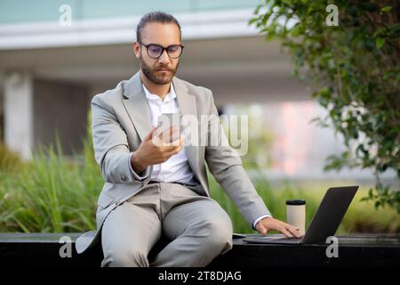 Young professional businessman in suit multitasking with smartphone and laptop outdoors Stock Photo