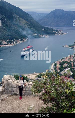 A tourist takes a selfie at Kotor Fortress with a backdrop  of a cruise ship in the Bay of Kotor, Montenegro, 2023 Stock Photo