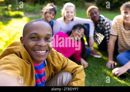 Portrait of happy diverse teenage friends taking selfie and sitting on grass in park Stock Photo