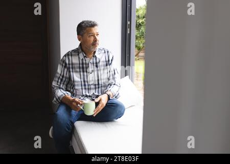 Thoughtful senior biracial man drinking coffee at window at home Stock Photo