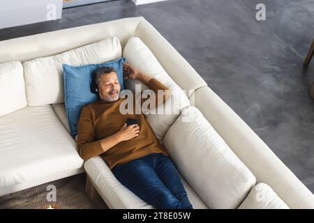 Happy senior biracial man lying on couch with headphones and using smartphone at home Stock Photo