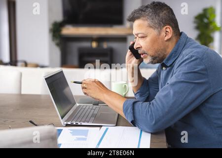 Busy senior biracial man talking on smartphone and working on laptop at home Stock Photo