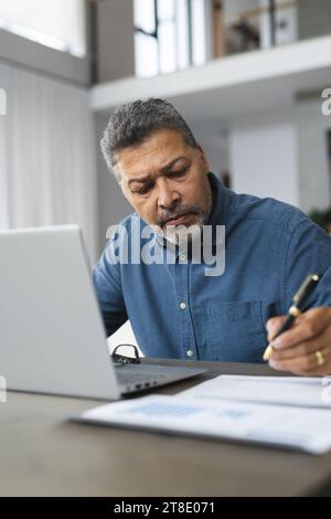 Busy senior biracial man taking notes and working on laptop at home Stock Photo