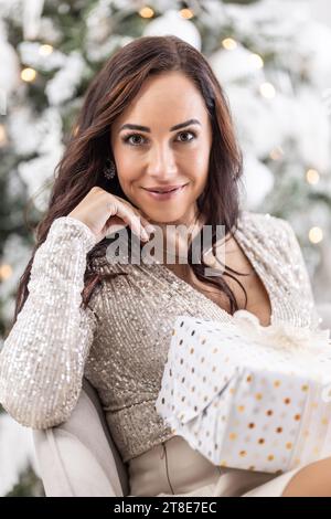 Beautiful woman holds Christmas gift with an Xmas tree in the background. Stock Photo