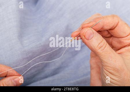 Close up photo extreme horizontal, hands mature adult Caucasian woman, threading needle. Preparing sewing material to repair a blue shirt doing tailol Stock Photo