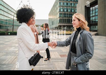 Friendly businesswoman greeting and handshake of new staff member. Young female business people on corporate sales department standing outdoors. Mature woman welcoming a new job candidate at workplace. High quality photo Stock Photo