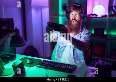 Young redhead man streamer playing video game using smartphone at gaming room Stock Photo