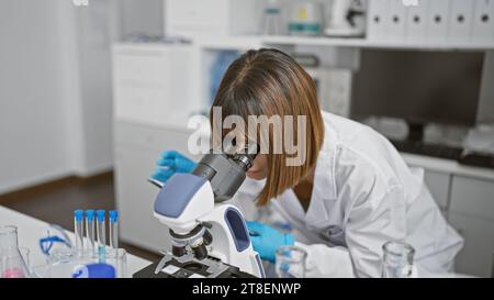 Relaxed yet focused, a young, beautiful hispanic woman scientist working diligently indoors at the lab, using a microscope. she's deeply engrossed in Stock Photo