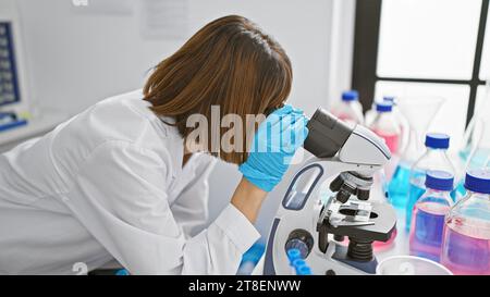 Relaxed yet focused, a young, beautiful hispanic woman scientist working diligently indoors at the lab, using a microscope. she's deeply engrossed in Stock Photo