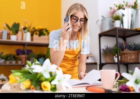 Young blonde woman florist talking on smartphone reading book at flower shop Stock Photo