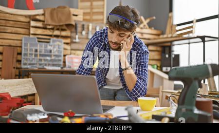 Handsome young arab man engrossed in serious carpentry business conversation using smartphone, while surfing the internet on his laptop at the woodwor Stock Photo