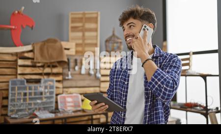 Handsome young arab man carpenter talking on smartphone, beaming with smiling face, engrossed in woodworking on touchpad indoors at his carpentry work Stock Photo