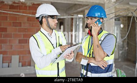 Two men builders talking on smartphone reading document at construction site Stock Photo