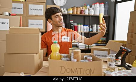 Smiling young hispanic man volunteer taking an altruistic selfie with a cellphone in a cheerful charity center, making memories while working indoors Stock Photo