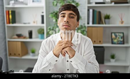 Handsome, young hispanic man sits at office table, embodying confidence. this serious business worker's focused gaze screams success as he takes a bre Stock Photo