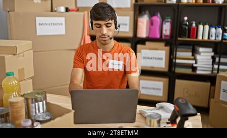 Hardworking young hispanic man volunteering at a charity center, immersed in his helpdesk tasks, wearing headphones and operating laptop Stock Photo