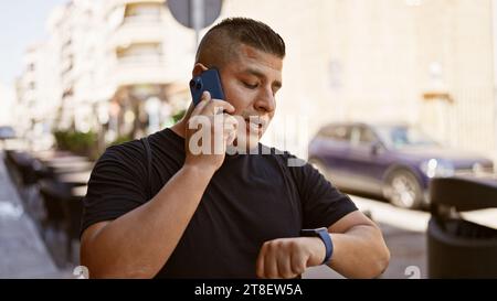 Casual latin man, talking seriously on his smartphone, checks the time on his wristwatch while standing under the sunny urban city outdoors. Stock Photo