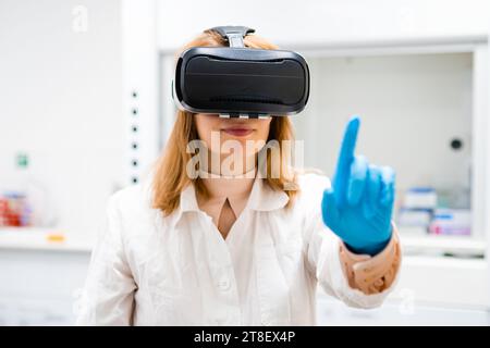 Portrait of young scientist in VR glasses moves finger imitating touch. Curious blonde woman in rubber gloves tries virtual reality in modern laboratory Stock Photo