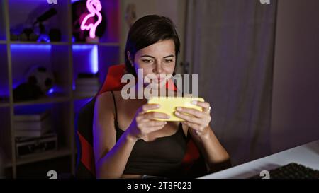 Confident young hispanic woman, streams smiling and playing video games in home gaming room, using phone Stock Photo