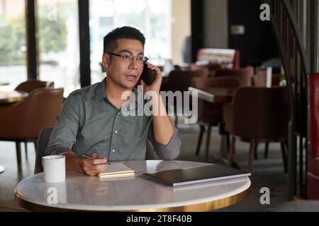 Entrepreneur sitting at cafe table, talking on phone with coworker and taking notes in planner Stock Photo
