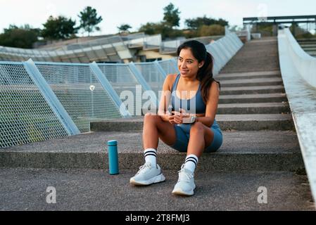 A smiling Latin American woman takes a break on stadium steps, holding her phone and sitting next to a water bottle Stock Photo