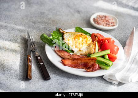 A balanced breakfast arrangement on a white plate with fried egg, crispy bacon, fresh spinach leaves, sliced cucumber and wedged tomatoes served with Stock Photo