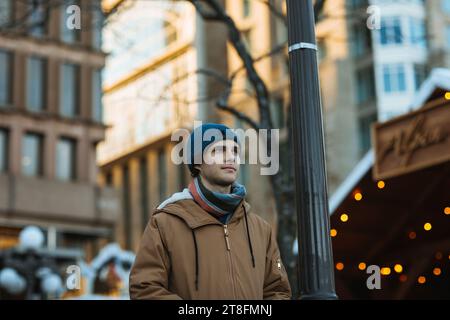 A young adult male in a casual winter outfit stands beside a lamppost on a city street, looking thoughtfully into the distance in Quebec, Canada Stock Photo