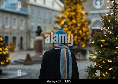 A person in a winter hat stands facing a twinkling Christmas tree in an urban square, creating a cozy holiday atmosphere in Quebec, Canada Stock Photo