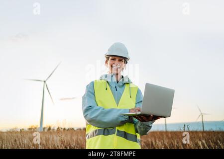 Female engineer looking at camera in safety gear standing with a laptop in a field at sunset, with wind turbines in the background, representing renew Stock Photo