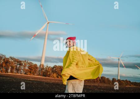 A cheerful individual wearing a yellow raincoat and a pink beanie smiles in a field with wind turbines in the background under a beautiful sunset sky. Stock Photo