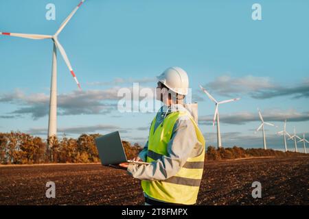 An engineer in a high-visibility vest and safety helmet analyzes data on a laptop with wind turbines in the background during sunset. Stock Photo