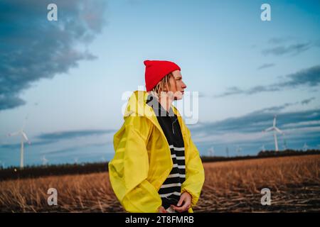 A pensive young male in a bright yellow raincoat and red beanie stands in a field with wind turbines under a vast sky at dusk. Stock Photo