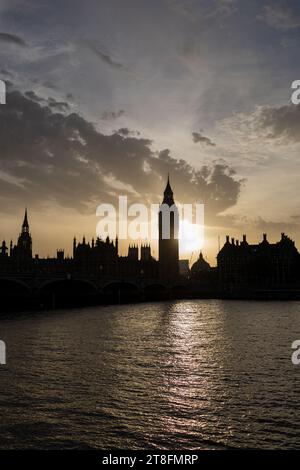The iconic Big Ben and Houses of Parliament stand silhouetted against a dramatic sunset sky in London, with reflections on the Thames River. Stock Photo