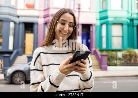 Cheerful young female in casual attire enjoys her smartphone on a vibrant, colored backdrop in London. Stock Photo