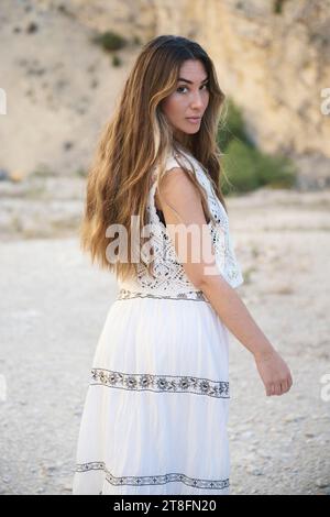 A young woman in a white sleeveless dress with decorative patterns outdoors looking at camera with her long brown hair down her back Stock Photo