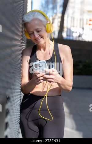 An active senior woman with headphones uses a smartphone while taking a break from her workout on a sunny day. Stock Photo