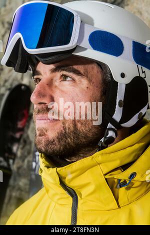 A close-up of a bearded male skier wearing a yellow jacket, helmet, and goggles, gazing into the distance with a thoughtful expression. Stock Photo