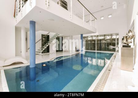 Swimming pool with large window and staircase leading to upper floor in luxurious modern house with white walls Stock Photo