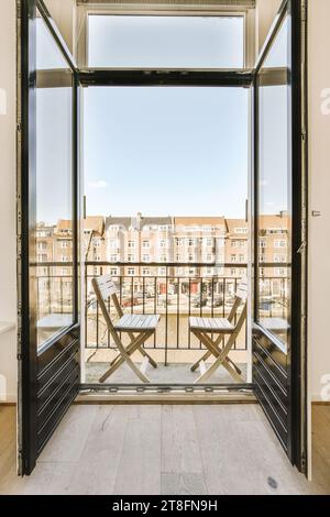 Open glass doors leading to balcony with empty chairs overlooking residential buildings against blue sky in city Stock Photo