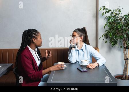 Two young business woman colleagues taking a break in nearby cafeteria drinking coffee talking about private life, to get know each other better. Staf Stock Photo