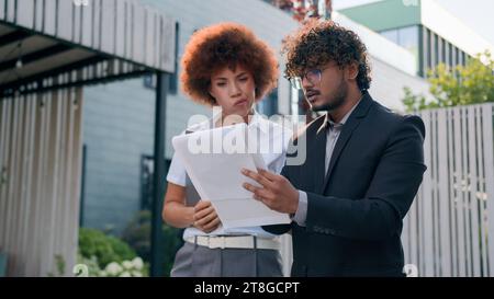 Two confused anxious diverse coworkers walking in city check papers bad result talking problem Indian businessman business talk with African Stock Photo