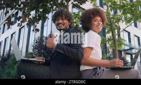Two business partners sit outdoors on bench multiracial colleagues diverse co-workers in city working with computers laptops showing thumb up Stock Photo