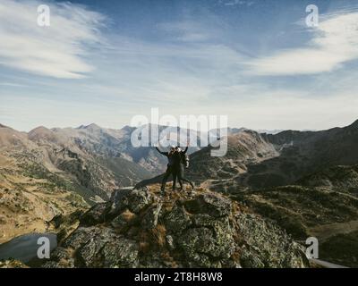 A man and a woman stand atop a mountain peak, looking out over the vast expanse of majestic mountains Stock Photo