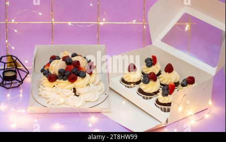 Beautiful cake with whipped cream and fresh berries and cupcakes in the gift box on festive christmas background. Bokeh, string lights Stock Photo