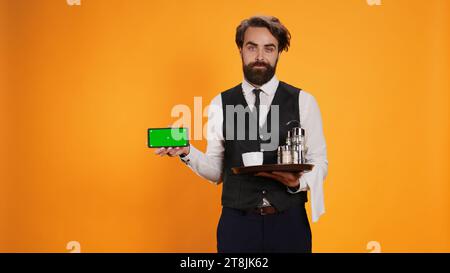 Luxurious worker points at greenscreen on smartphone, presenting blank copyspace display on mobile device in studio. Skilled employee holding phone with empty mockup screen on camera. Stock Photo