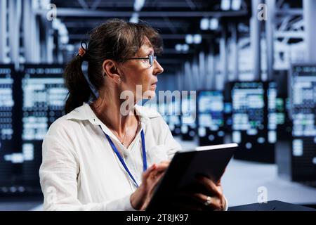 Elderly efficient admin updating data center units, writing intricate binary code scripts on tablet, using programming to fortify cybersecurity in technically advanced server farm Stock Photo