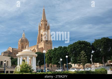 View of the church Madonna ta Loreto, in Ghajnsielem, Gozo. The church tower rises high above the town and the trees. In the background blue sky with Stock Photo